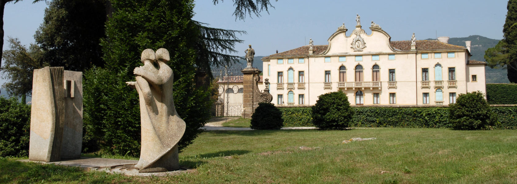 Scultura in pietra di due figure in primo piano in esposizione nel Parco della Villa di Montruglio, con sfondo della Facciata.