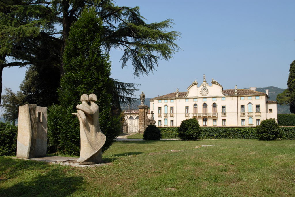 Scultura in pietra di due figure in primo piano in esposizione nel Parco della Villa di Montruglio, con sfondo della Barchessa.