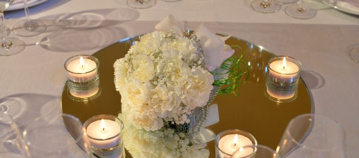 Dettagli di mise en place, con bouquet e candeline, sopra ad un tavolo bianco, circondato da bicchieri e piatti.