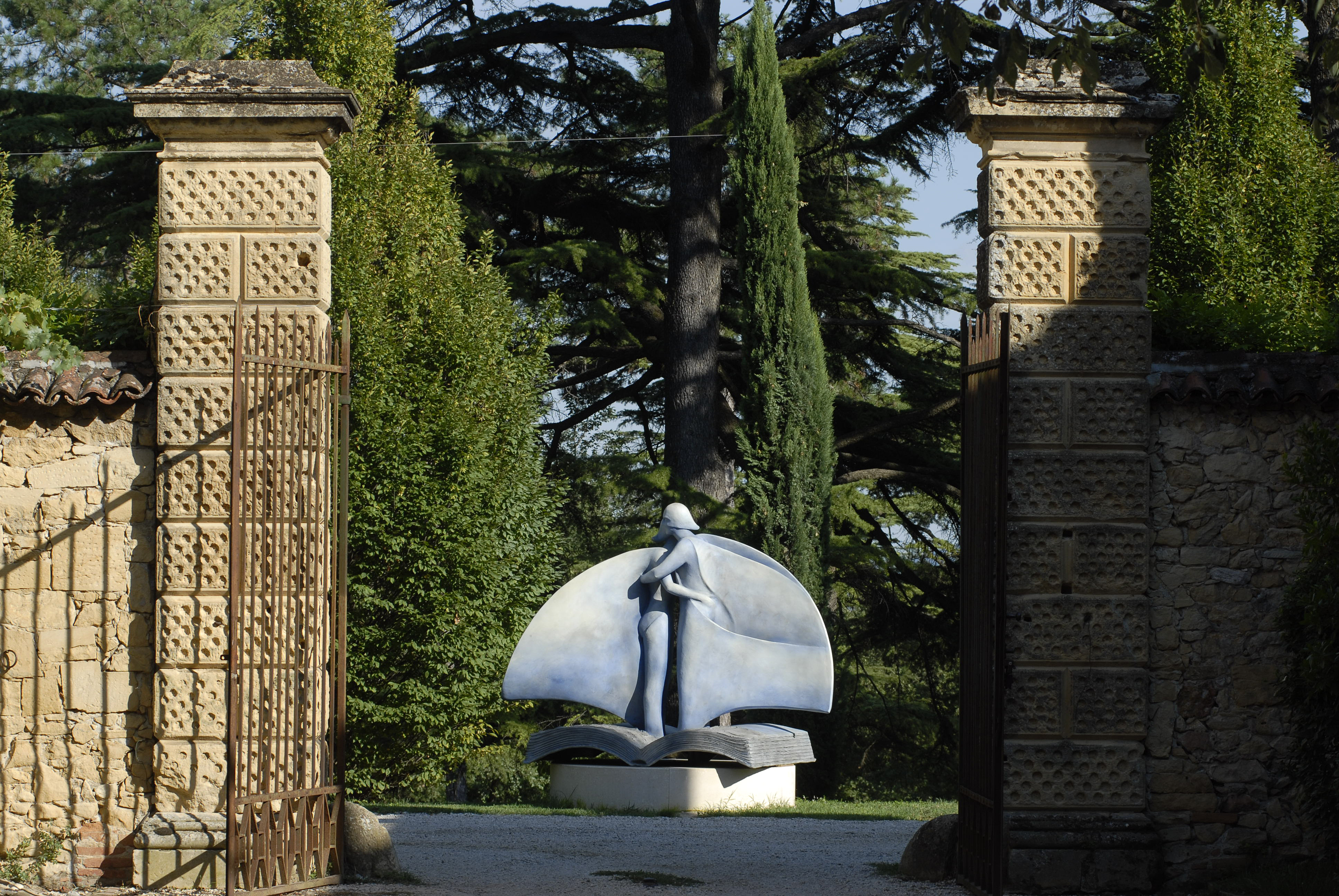 Vista dal cancello dell'entrata di una scultura in pietra di due figure abbracciate su di un piedistallo a forma di libro, nel Parco della Villa di Montruglio. Sullo sfondo degli alberi.
