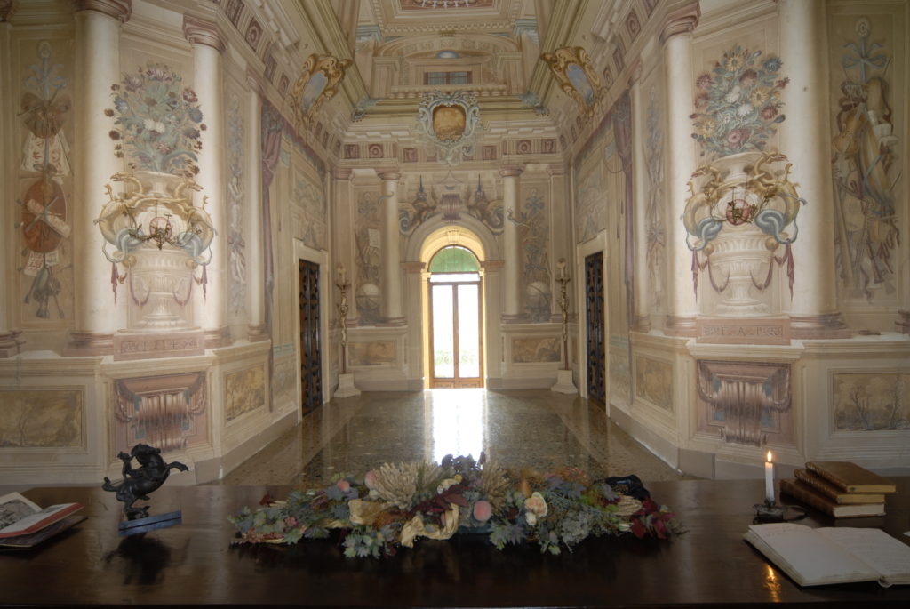 Vista interna della Villa di Montruglio con pareti affrescate, una porta illuminata sul fondo, due laterali e in primo piano una scrivania con dettagli floreali.