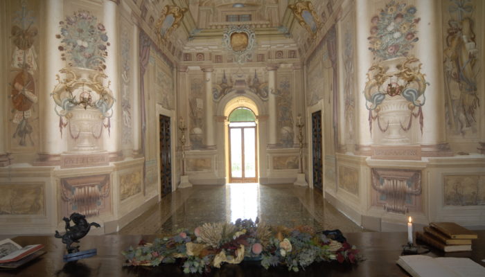 Vista interna delle pareti affrescate della Villa di Montruglio, con delle porte speculari ed una finestra luminosa sul fondo.