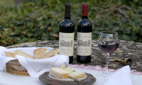 Visione di un bicchiere di vino e due bottiglie della Villa di Montruglio, con in primo piano un piatto di formaggi e un cestino di pane.