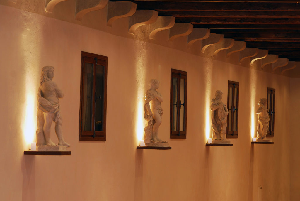 Piccole sculture statuarie poste vicine alla parete su piedistalli aerei, con illuminazione dal basso, nella Villa di Montruglio.