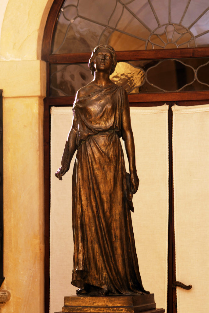 Scultura bronzea di una donna su piedistallo, con sfondo di un arco.